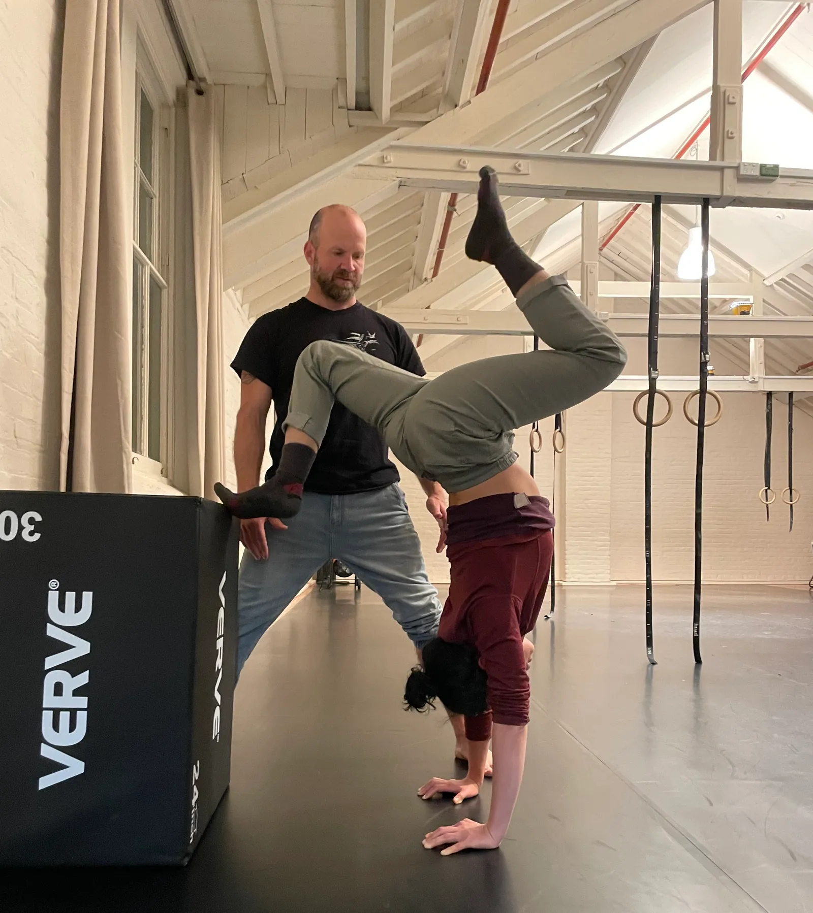 Scott spotting a student mid-handstand in the studio, rings hanging on the wall behind them