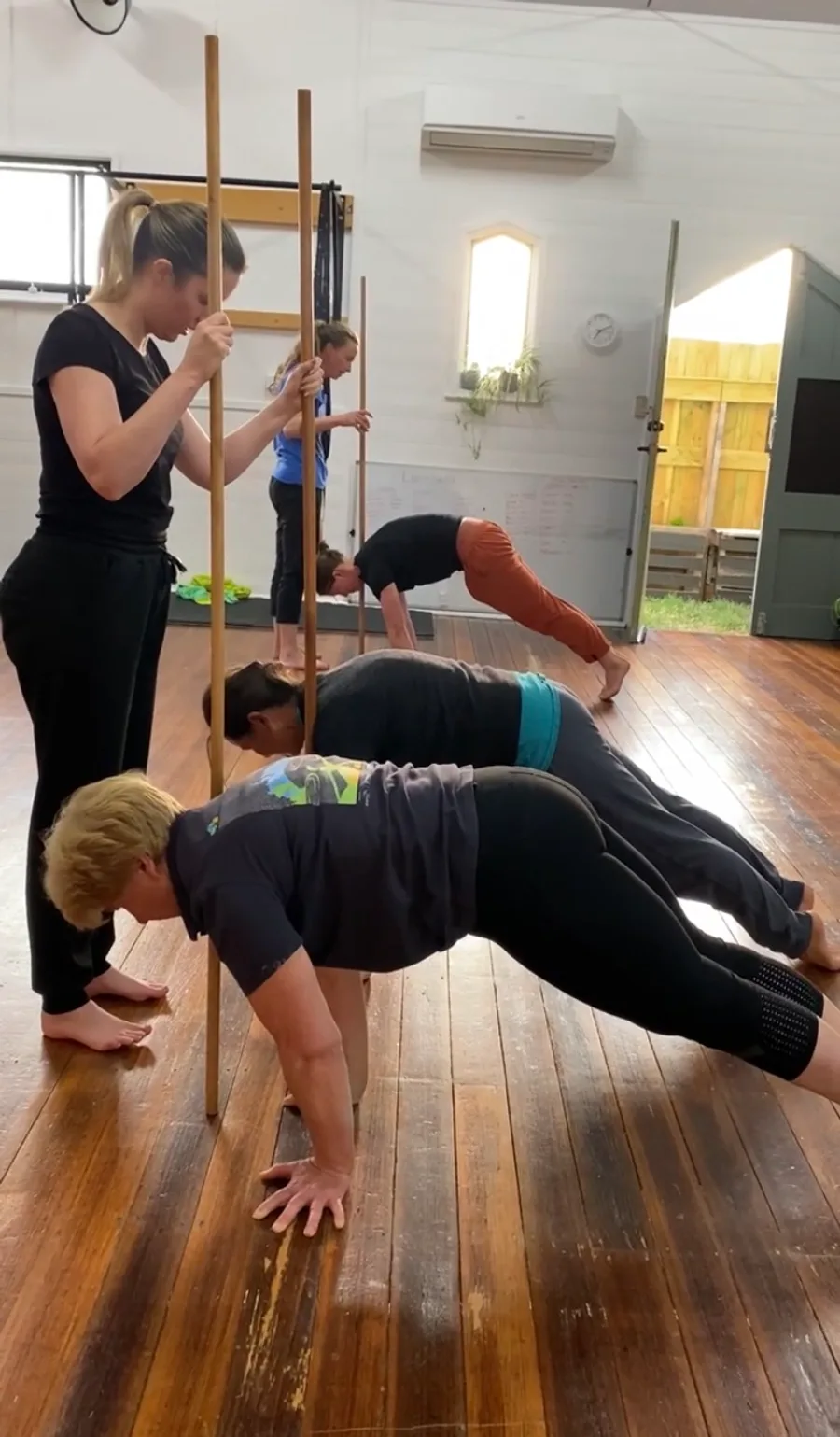 A small group holding a plank while a coach checks their alignment against a long wooden dowel