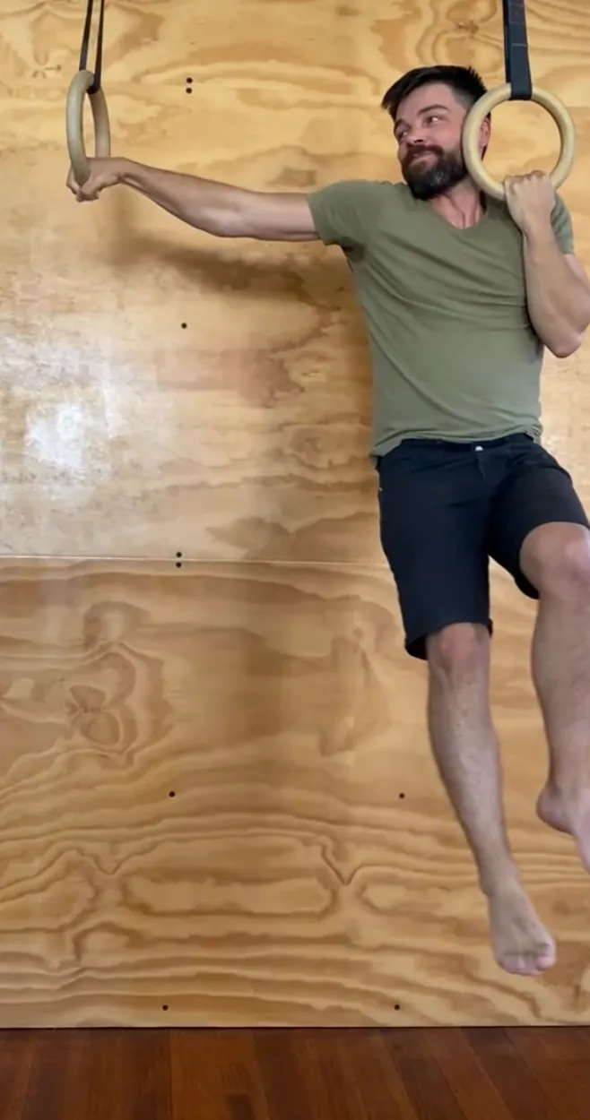A student hanging from the rings in an archer pull, one arm straight and one bent, feet clear of the floor in front of the plywood training wall