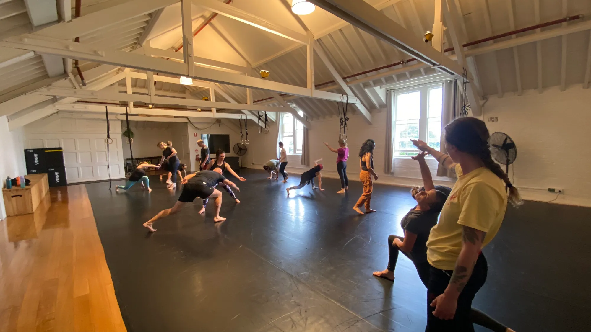 Inside the Totum studio: a large light-filled room under a white-beamed ceiling, rings hanging from the rafters, a class spread across the floor mid-movement